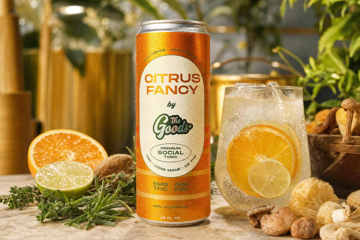 Orange "Citrus Fancy by The Goods" THC/CBD social tonic can beside an iced citrus drink, fresh fruit, herbs, and mushrooms.