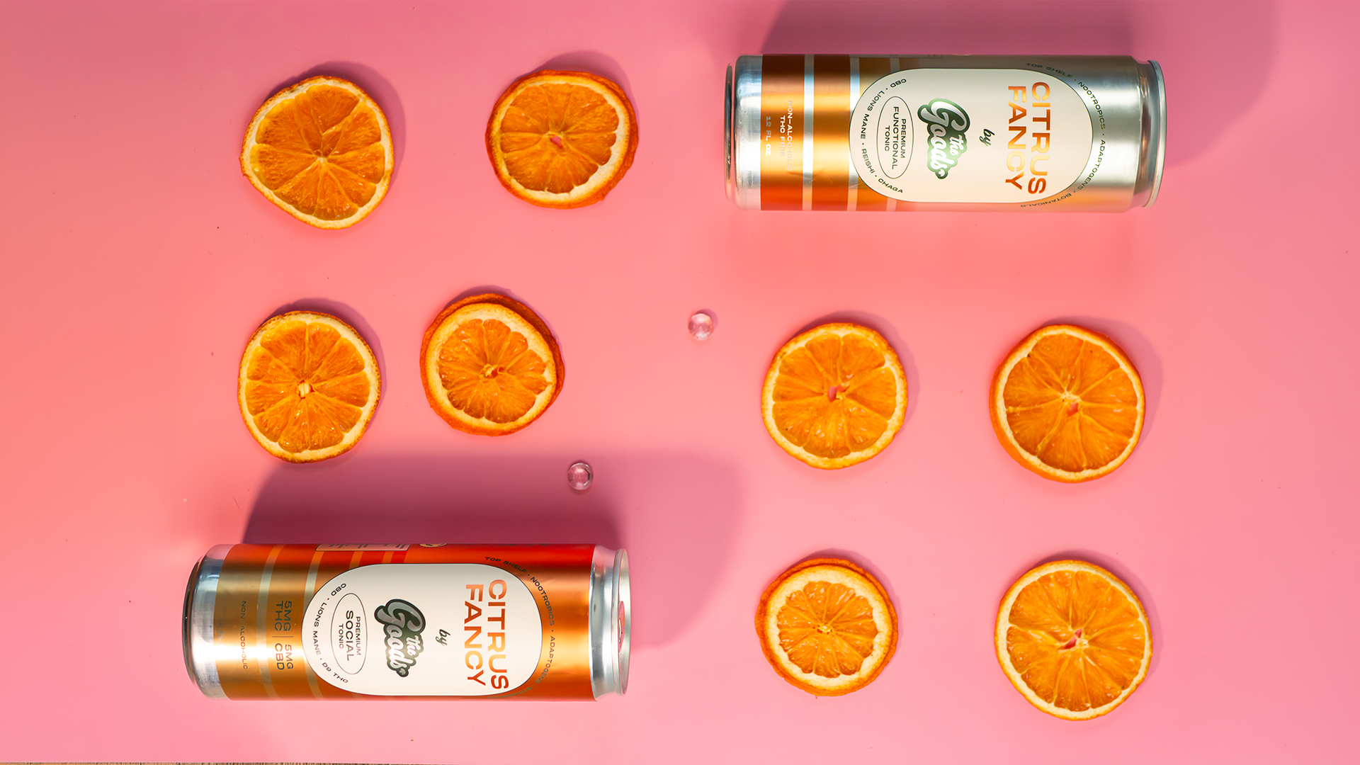 two cans of The Goods Citrus Fancy functional tonic in front of a pink background surrounded by orange slices